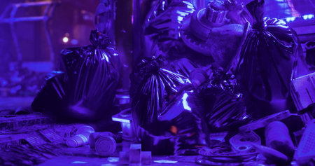 Black garbage bags accumulate in a dimly lit urban setting, surrounded by scattered debris and lit...
