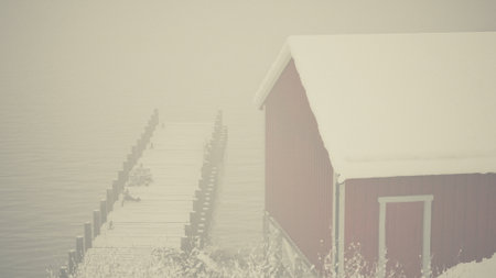 A red barn stands alongside a body of water covered in snow, with an old wooden pier stretching into the cold sea.の写真素材