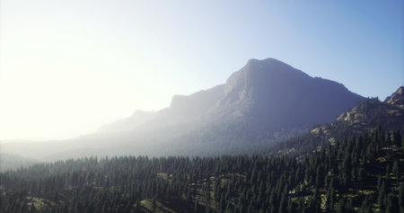 Vast mountain range bathed in golden morning light rises above a dense forest, creating a peaceful...
