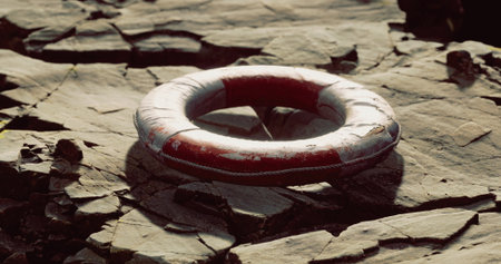A red lifebuoy lies abandoned on a rocky shoreline, surrounded by various stones. The sun casts...