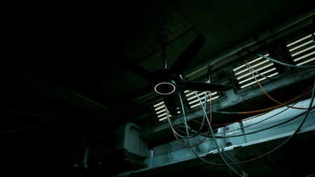 In a dim, shadowy space, an old ceiling fan slowly turns, its blades cutting through the still air. Tangled wires dangle from above, hinting at a forgotten project in this neglected area.の写真素材