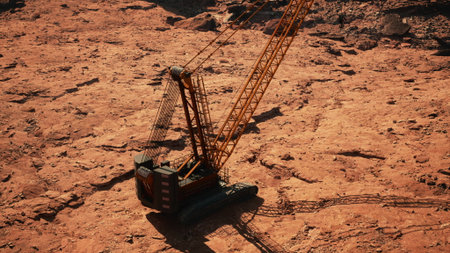 A large yellow crane stands on dry, reddish terrain, its shadow stretching across the ground. The...