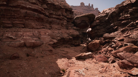 A rugged canyon path stretches through stunning red rock formations under the bright midday sun. Shadows dance on the terrain, while the towering cliffs rise dramatically around.の写真素材