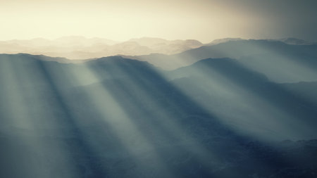 In the fading light of dusk, sun rays break through a veil of clouds, casting an ethereal glow over layered mountain ranges, creating a tranquil atmosphere filled with depth and beauty.の写真素材