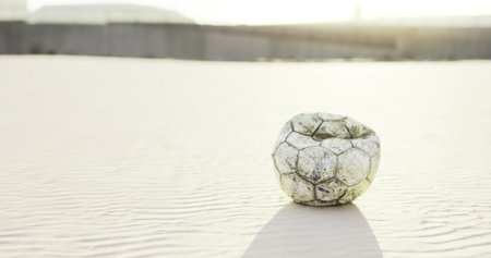 A weathered soccer ball sits alone on the soft sandy surface of a beach. The sun sets in the background, casting a warm glow over the scene, evoking feelings of nostalgia and play.の写真素材