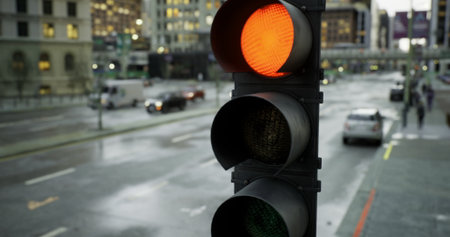 A traffic light shows an orange signal in a city setting, while raindrops create reflections on the wet pavement. People navigate the street as vehicles pass by, highlighting urban life.の写真素材