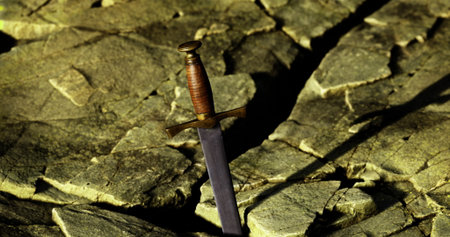 A sword stands prominently embedded in a crack of jagged rock, illuminated by warm light. The contrast of the metal against the stone creates a striking visual.の写真素材