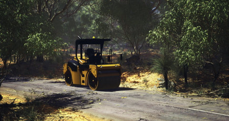 During a sunny day, a road roller works diligently on a dirt road surrounded by lush trees and...