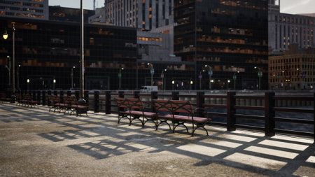 Benches line a serene riverside path as shadows stretch across the pavement. The scene captures a peaceful evening, with buildings softly glowing in the background.の写真素材