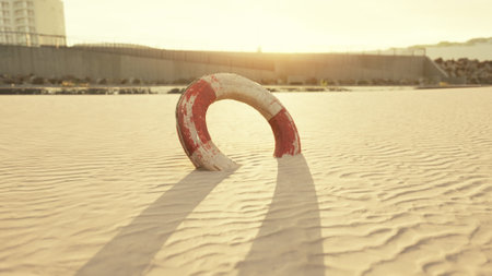 A lifebuoy rests on the soft, warm sand as the sun sets on the horizon. The golden light casts long shadows, enhancing the serene atmosphere by the waterfront.の写真素材