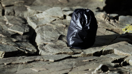 A black trash bag is placed on a rugged stone surface, surrounded by nature. The sunlight creates shadows, highlighting the contrast of the bag against the rocky terrain.の写真素材