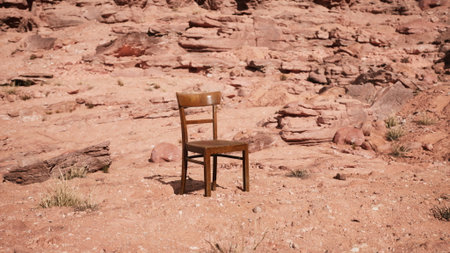 A solitary wooden chair stands out in a barren desert, surrounded by rugged red rock formations and dry soil. The stark contrast evokes feelings of isolation and serenity under the blue sky.の写真素材