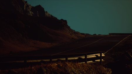 A winding road stretches through dark, rugged mountains under a night sky. The landscape features steep terrain, illuminated by an otherworldly glow, inviting adventure and reflection.の写真素材