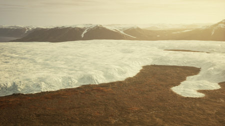The sun sets behind a vast glacier, casting golden light on a rugged terrain below. The icy expanse contrasts with earthy tones, creating a stunning and serene atmosphere in the wilderness.の写真素材