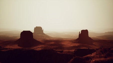 Golden light casts a warm glow over the rugged desert terrain as towering rock formations rise against a hazy sky. Shadows stretch across the ground, adding mystery to the scene.の写真素材