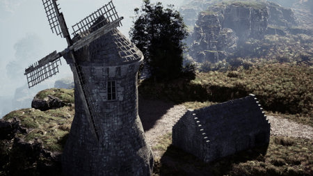 A scenic view features an old windmill adjacent to a quaint stone building, nestled in lush greenery against a backdrop of rocky cliffs. The atmosphere is serene and foggy.の写真素材