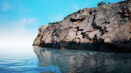 A serene view of a rocky shoreline meets tranquil ocean waters. The sky is bright and blue, casting reflections on the gentle waves. Natureâs beauty is on full display in this peaceful scene.の写真素材