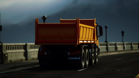 A bright orange dump truck navigates down a bridge as the sun sets, casting shadows against the gray clouds. Street lamps line the path as the day transitions to evening.の写真素材