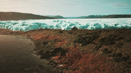 The sun rises over the icy glaciers in Greenland, reflecting light on the dark rocky shore. The serene landscape combines textures of ice and stone, creating a stunning natural scene.の写真素材