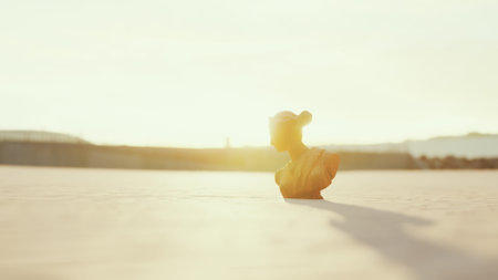 A small sculpture stands alone on a flat surface as the sun sets, casting warm golden light. The silhouette stretches, creating an artistic shadow while the scene radiates calmness.の写真素材