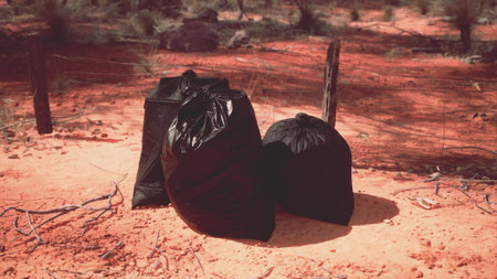 Three black litter bags are placed on the dry red earth. Sparse vegetation surrounds them under a bright sunny sky, highlighting the contrast of waste in a natural setting.の写真素材