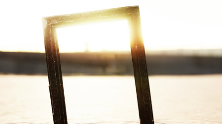 A vibrant sunset casts golden rays through an empty frame resting on warm sand. Waves gently lap at the shore, creating a serene atmosphere as distant figures stroll along the beach.の写真素材