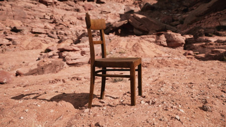 A wooden chair rests solitary on the dry earth amidst rugged rock formations. Sunlight casts shadows, creating a serene yet striking contrast against the natural beauty of the desert.の写真素材