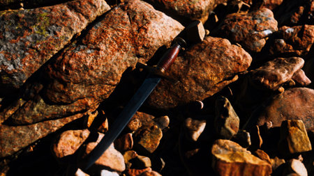A rustic dagger with a wooden handle lies partially hidden among colorful stones in a rocky terrain. The warm glow of sunset casts a creative light over the scene, highlighting textures.の写真素材
