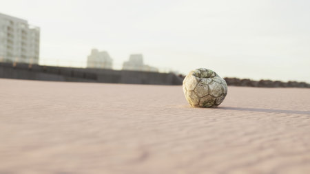 A weathered soccer ball sits alone on a sandy surface, surrounded by a soft glow from the sun. Nearby, tall buildings loom in the distance, hinting at an urban setting.の写真素材