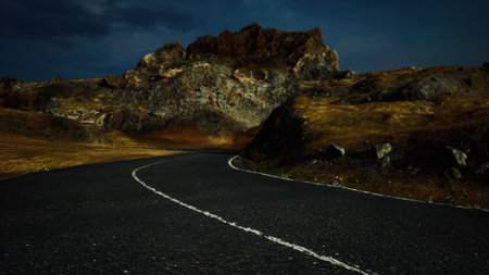 A winding road leads through a desolate landscape at dusk, framed by towering rugged mountains. The sky is darkening, creating an intense and atmospheric scene filled with mystery.の写真素材