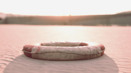 A lifebuoy lies abandoned on a sandy beach, illuminated by the soft glow of the setting sun. Waves gently lap the shore, creating a serene atmosphere.の写真素材