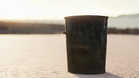 A weathered trash can rests quietly on a beach as the sun sets, casting warm golden hues on the sand and creating a serene atmosphere around it.の写真素材