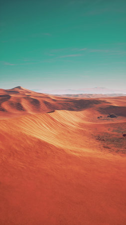 A vast desert stretches across the horizon, showcasing rolling sand dunes in warm shades of orange and yellow, with a clear blue sky above and distant mountains on the edge of the scene.の写真素材