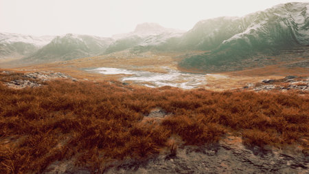 Sunlight gently illuminates a tranquil valley filled with golden grass. Snow capped mountains rise in the background, and a small lake reflects the soft morning light.の写真素材
