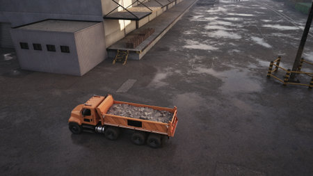 An orange dump truck drives across a wet construction site in cloudy weather. The ground is slick with puddles, revealing the busy atmosphere of ongoing work.の写真素材