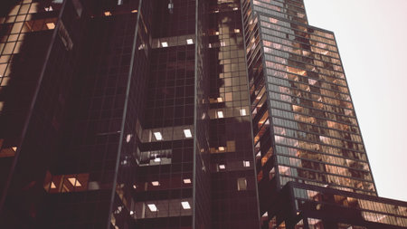 Tall buildings stretch towards the sky, their glass surfaces reflecting warm evening light. The urban environment shows hints of life within the multiple windows, creating a dynamic city vibe.の写真素材