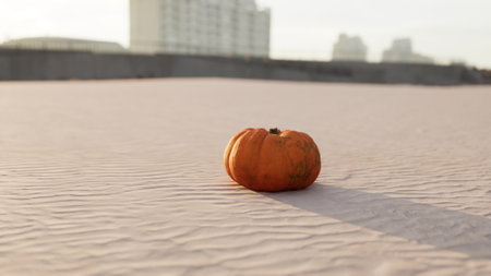 A vibrant orange pumpkin stands alone on smooth sand, surrounded by a serene urban landscape as the sun sets. Shadows stretch across the surface, adding warmth to the scene.の写真素材