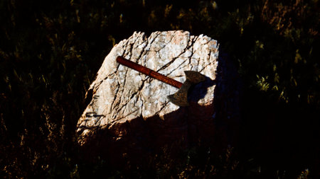 An old, rusty axe is positioned on a large rock under the soft glow of dusk. The surrounding field shows tall grasses and a hint of twilight, creating a serene atmosphere.の写真素材