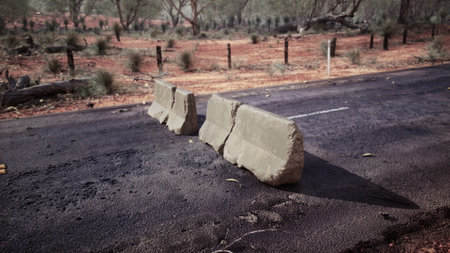 Two large concrete blocks obstruct a road in a vast outback setting, surrounded by dry vegetation and reddish earth. The bright sunlight emphasizes the desolate beauty of the scene.の写真素材