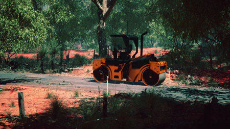 A road roller stands still on a newly paved road surrounded by lush greenery and reddish earth....