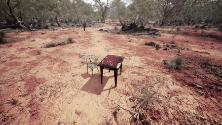 A solitary table and chair sit on cracked, dry earth, surrounded by sparse vegetation and distant trees. The abandoned scene highlights a stark contrast between furniture and the rugged, arid terrain.の写真素材