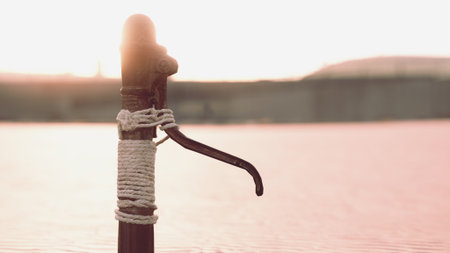 A weathered water pump is tied with rope, casting a long shadow over the calm lake. The sun sets in...