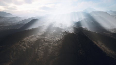 Sunlight streams through clouds, casting radiant beams over a rugged mountain terrain. The atmosphere is serene, creating a mystical feel to the landscape at dawn.の写真素材