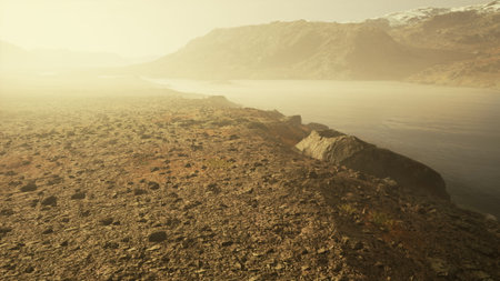 A serene scene unfolds at dawn where the river gently hugs the rocky shore. Mist rises, creating an ethereal atmosphere as the mountains loom in the background, partially obscured by fog.の写真素材