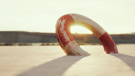 A lifebuoy leans against the warm sand at sunset, casting long shadows in the soft light. The colors of the sunset reflect on the surface, creating a serene and peaceful scene by the beach.の写真素材