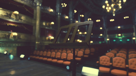 The stage awaits as a music stand stands ready in a beautiful theater. Plush orange seats line the space, illuminated by soft lights in anticipation of the upcoming show.の写真素材