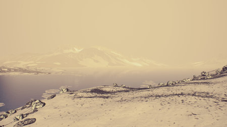 A serene winter scene features a rocky hillside leading to a calm body of water. The soft golden light creates a peaceful atmosphere, enhancing the beauty of the surrounding mountains.の写真素材