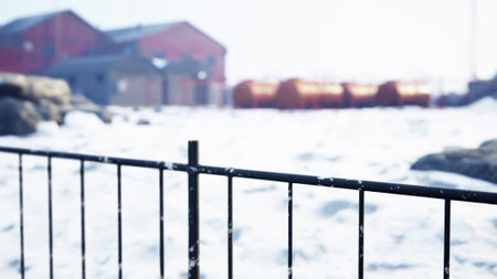 An icy landscape captures a winter scene along the coast. A black fence outlines the foreground while snow covers the ground. In the distance, red buildings emerge against a pale sky.の写真素材