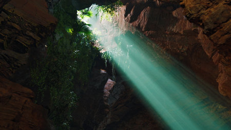 Bright shafts of sunlight penetrate a cave, illuminating vibrant green plants and rocky textures....