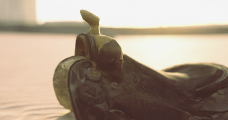 A beautifully crafted cowboy saddle rests on the sandy shore, glistening in the sunset light. The tranquil atmosphere captures the essence of rugged outdoor adventures and western heritage.の写真素材